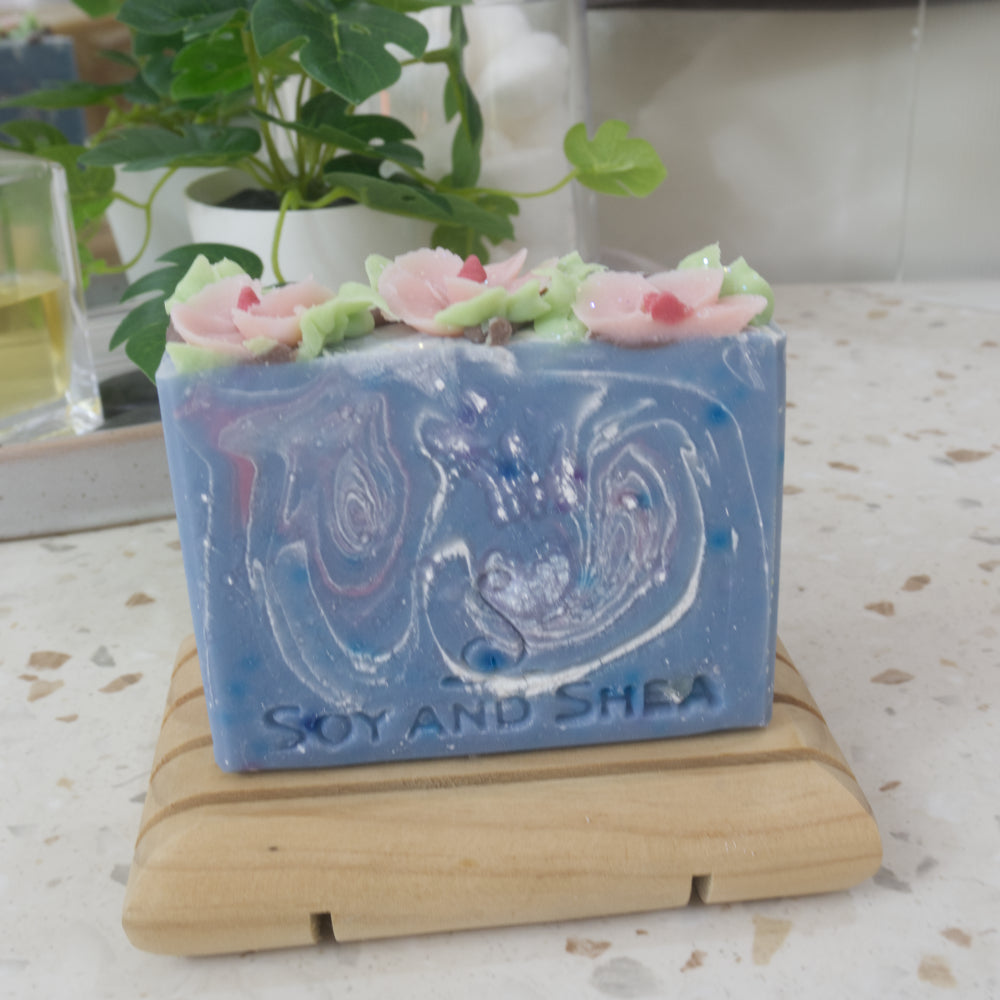 A bar of soap stands on a wooden soap dish on a bathroom counter top.  The soap has a blue base colour with swirls of blue and pink.  The top has pink flowers and green leaves piped with soap.