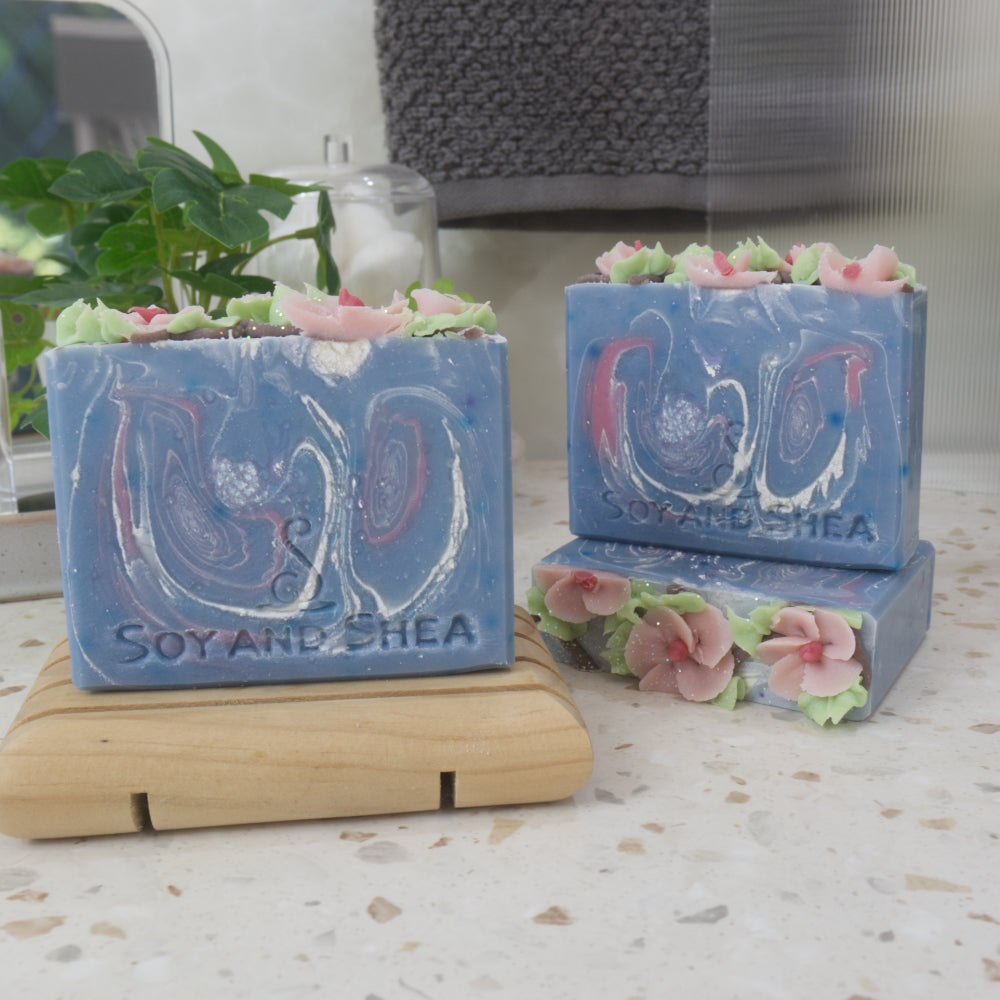 A selection of soaps are presented on a bathroom countertop along with a plant, hanging towel and jar with cotton.   The soap to the front left stands on a wooden soap dish and to the right and set back, a soap lays flat showing the decorative soap top.  A third soap stands upright on this soap.   All the soaps have a base blue colour with various white and pink swirls.  The tops are decorated with pink flowers and green leaves piped with soap.