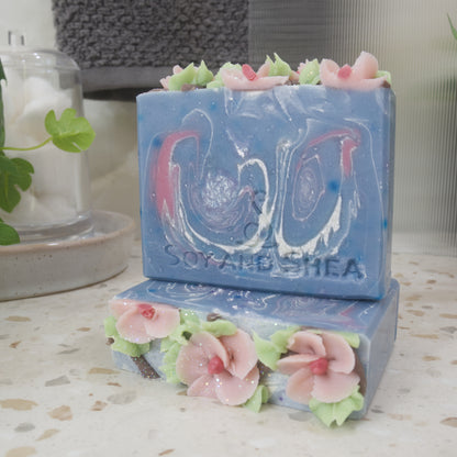 A soap lays flat on a bathroom countertop revealing a decorative top of pink flowers with pale green leaves all piped with soap.  Anoither soap stands upright on this soap showing a soap with a base colour of blue with white and pink swirls of colour.