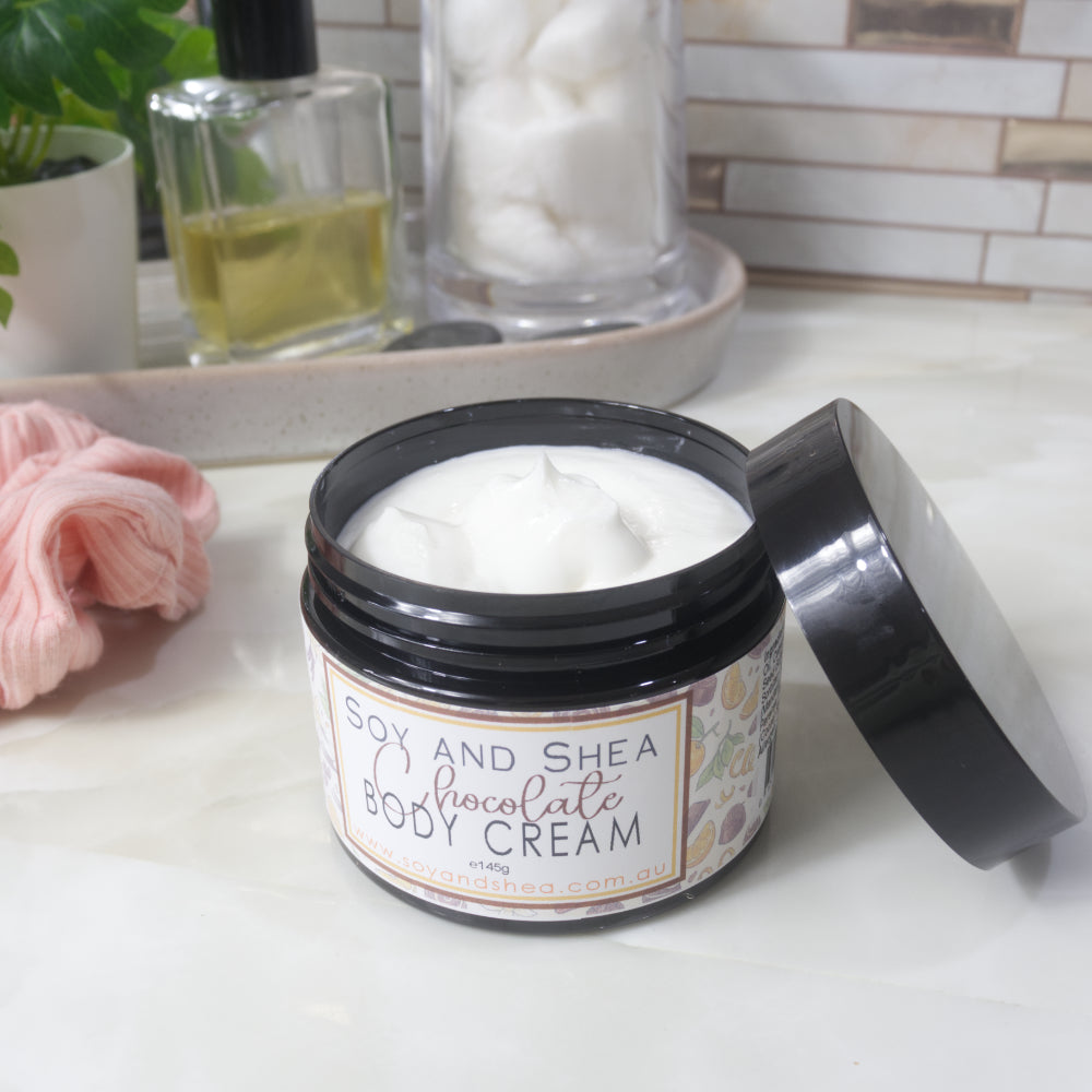 A black jar of Chocolate Body cream sits on a bathroom countertop with the lid removed and resting against the side of the jar.  The cream inside is white