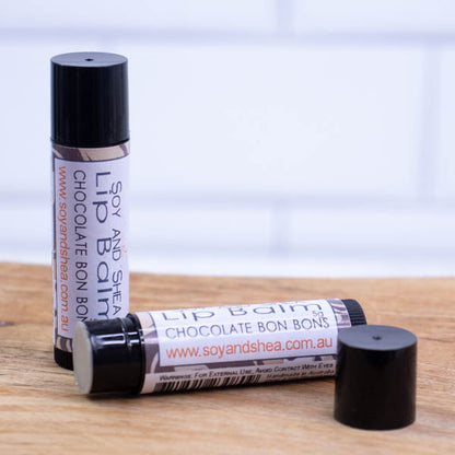 Two black tubes of lip balm are displayed on a wooden surface with blurred tiled background.  One tube stands on its end while the other lays flat in front with the cap removed revealing a cream balm.  The label has a swirling pattern in shades of brown.