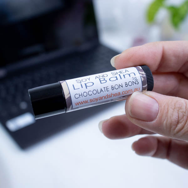 A hand holds a black tube of lip balm against a blurred background with an open laptop computer on a white surface.  The label has text LIP BALM CHOCOLATE BON BONS on a white background with a border of swirled brown.