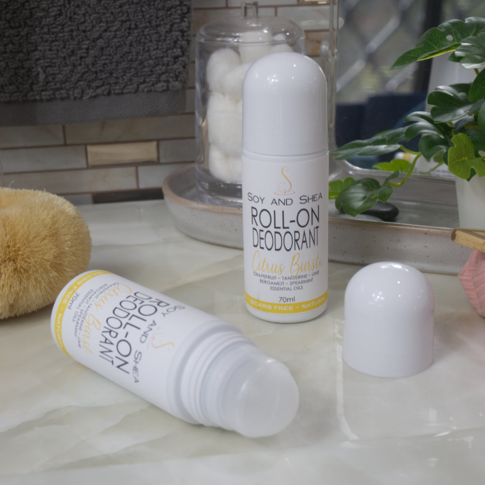 Two bottles of Roll-On Deodorant are displayed on a bathroom counter top with a jar of cotton balls, body brush and greenery.  One bottle stands upright with a domed cap, while another lays flay with the cap removed showing a ball roller in the top