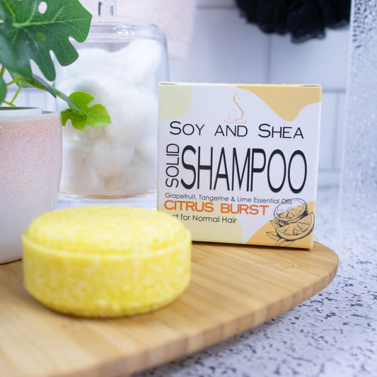 On a wooden tray sits a round yellow Shampoo bar and its cardboard box which is white with an abstract yellow design.  Next to it is a potted plant and a jar of cotton balls against a white tile background.