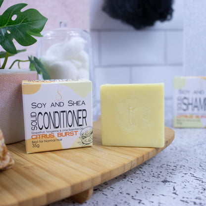 A wooden tray sits on a bathroom counter with a potted plant and jar of cotton balls against a blurred tiled background. Towards the front of the tray stands a white box with yellow abstract pattern with text SOLID CONDITIONER and next to it stands a square yellow bar of conditioner.