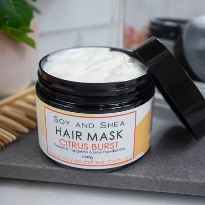 A black jar with the lid resting on its side revealing a white cream inside, sits on a dark grey tile surface against a blurred background of a bathroom counter top. The jar features a white label with yellow abstract design with text HAIR MASK CITRUS BURST.