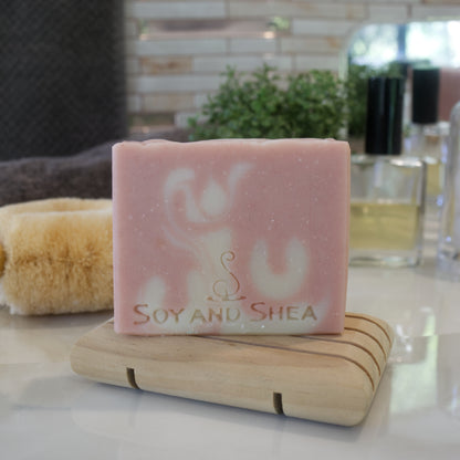 A pale pink soap bar with white swirls sits on a wooden soap dish surrounded by a grey towel, plant and bottle of perfume in front of a mirror.