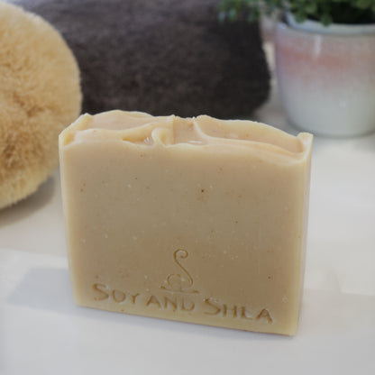 A pale yellow soap with flecks of dark yellow sits on a white bench in front of a grey towel and potted plant.