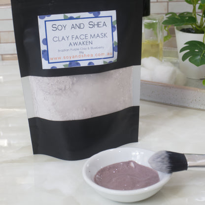 Soy and Shea Awaken Clay Face Mask Package on a bathroom counter.  In front of the package is a small bowl of hydrated mask along with a makeup brush 
