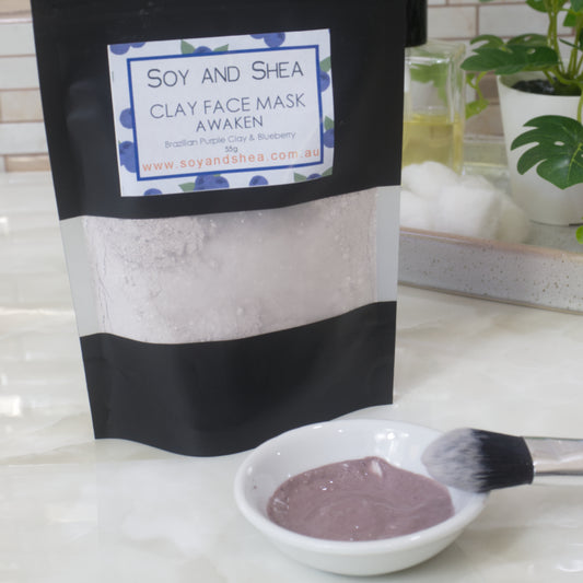 Soy and Shea Awaken Clay Face Mask Package on a bathroom counter.  In front of the package is a small bowl of hydrated mask along with a makeup brush 