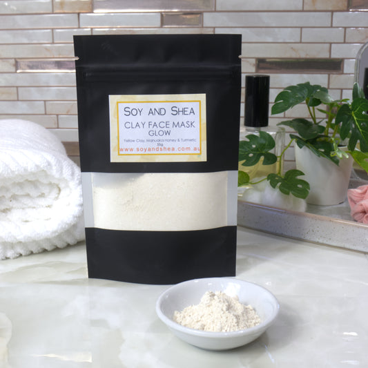 Soy and Shea Glow clay face mask packaging with a bowl of yellow powder on a bathroom counter.