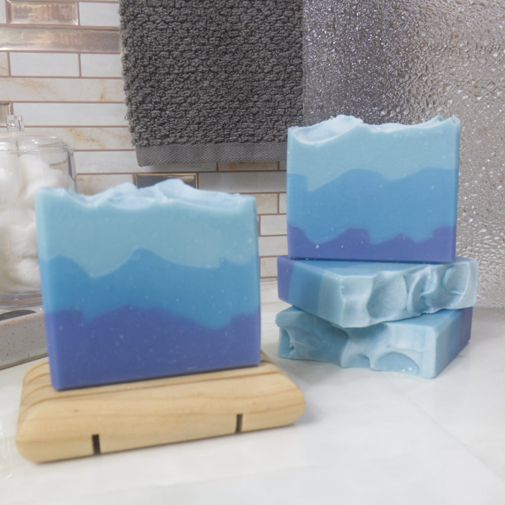 A selection of soap bars are presented on a bathroom countertop.  To the left is a bar of soap standing upright on a wooden soap dish.  The the right two soaps lay flat stacked upon each other with a third standing upright on them.  The soaps all have slight variations on pattern but all feature three layers of blue colour with dark blue at the bottom and light blue at the top.  The layers are uneven