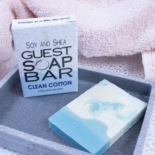 A grey stone soap dish sits next to a pink towel.  On the dish stands a white box with a border of light blue bubble foam across the bottom and text GUEST SOAP BAR. Next to it lies a small piece of soap which has a layer about the quarter of the soap in light blue with the rest in white with blue swirls.
