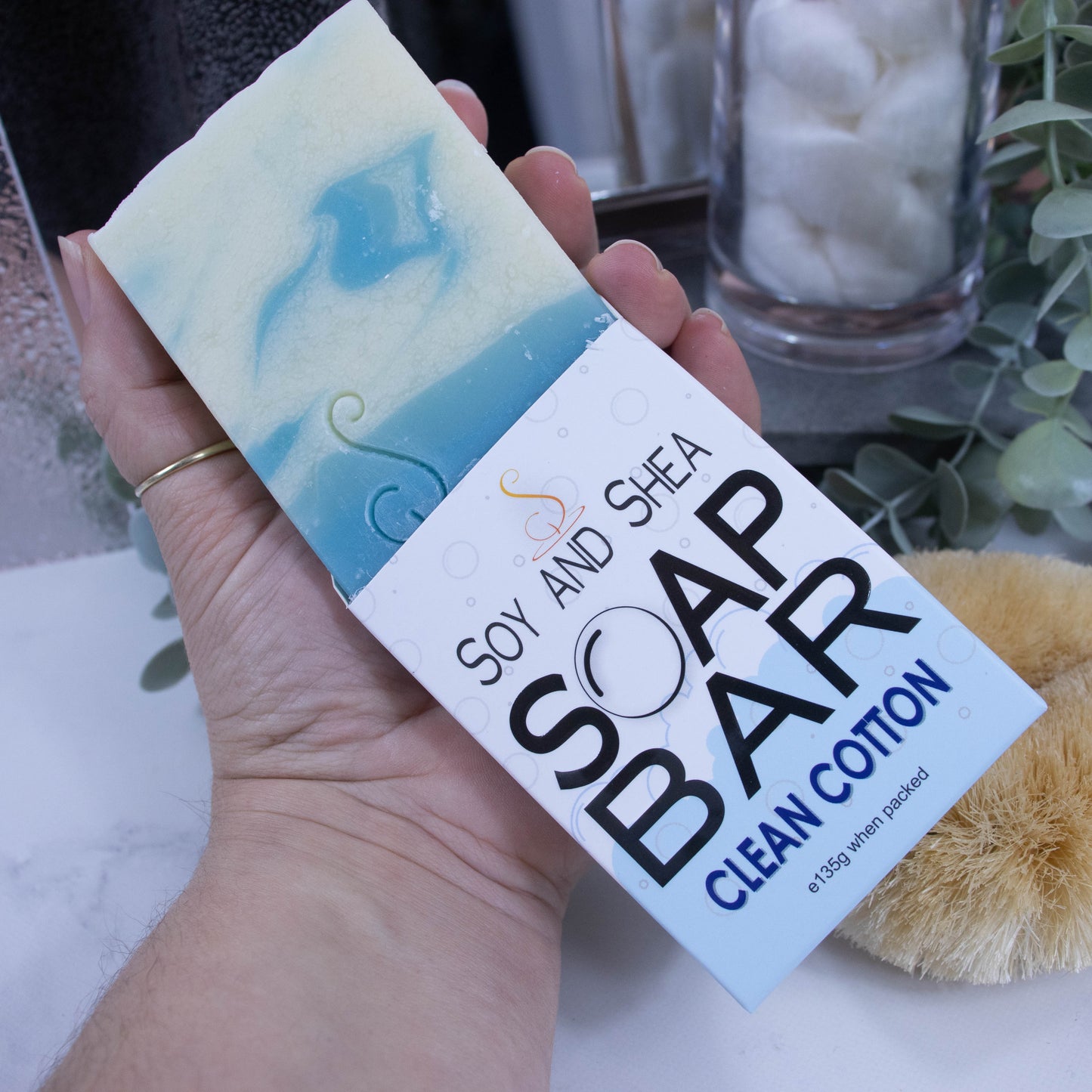 A hand is holding onto a box that has a bar of soap coming out the top. The lower half of the box has a pale blue bubble shaped border, and the top is white with grey bubbles. The box shows it is soap and the scent. The soap is rectangular in shape with a layered blue bottom and the top is white with blue swirls.  Behind is a bathroom countertop wiht mirror and cotton balls