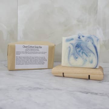 A white soap bar with blue swirls sits on a wooden stand with a brown paper package behind labeled 'Clean Cotton Soap Bar' on a marble background