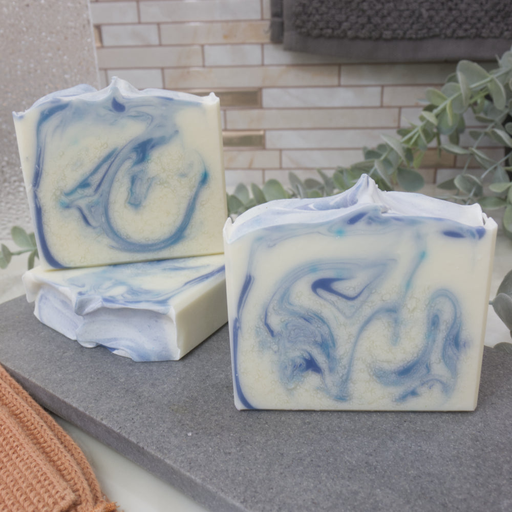 Three bars of soap with blue and white swirl patterns on a gray surface.  One soap is laying flat showing a textured top with another soap standing upright on it showing a swirl design.  Another soap sits in front with a different swirl design