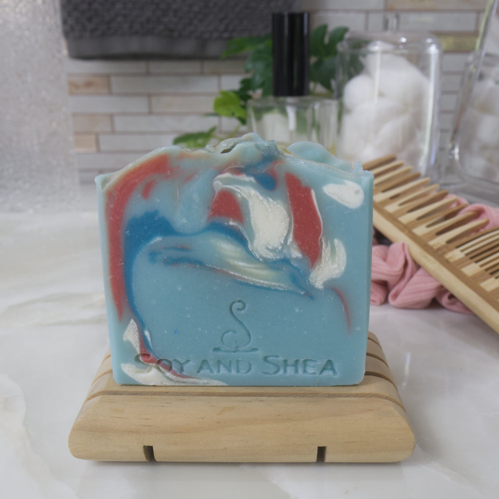 A light blue soap bar with Red, white and dark blue swirls on a wooden stand, branded 'Soy and Shea', in a bathroom setting.