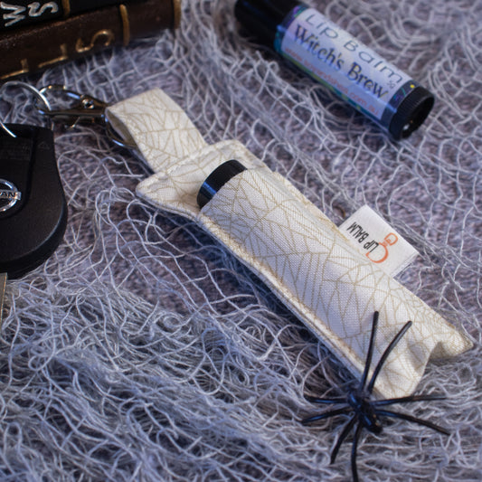 A Lip Balm holder lies on a grey mesh fabric attached to a car key with a lobster claw clasp.  Next to it is a lip balm.  The Clip Balm is cream in colour with a golden cobweb pattern