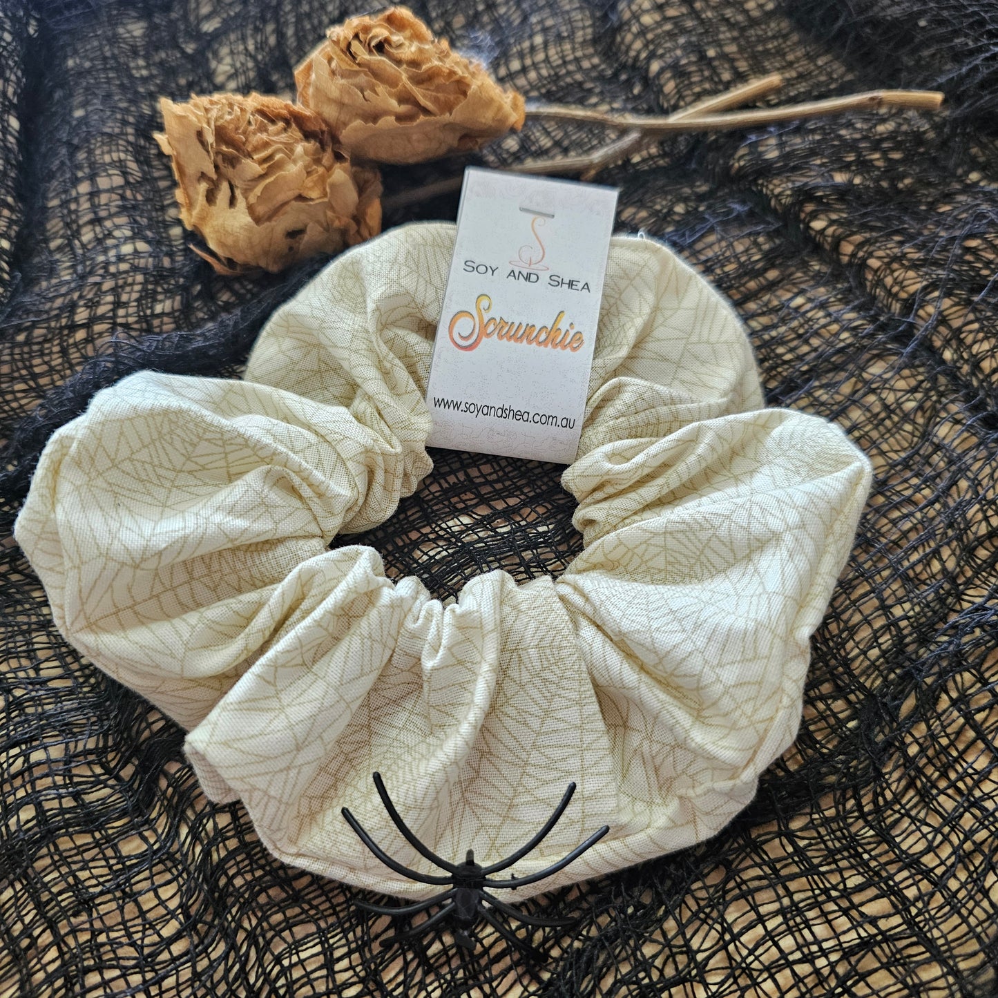 A beige scrunchie with a cobweb pattern in gold, displayed on a black mesh fabric with dried roses in the background.  