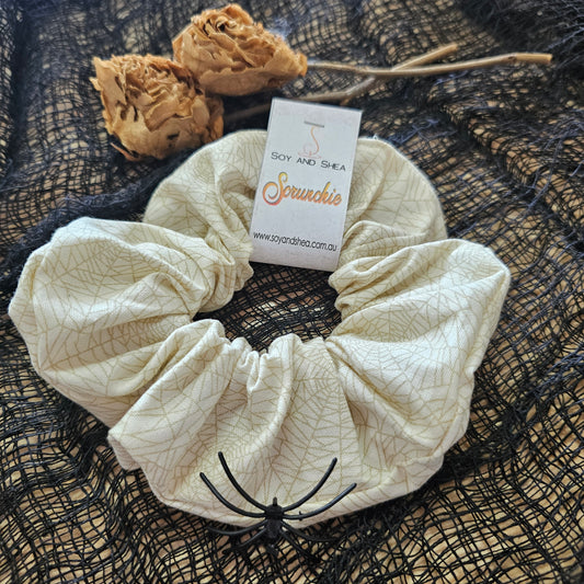 A beige scrunchie with a cobweb pattern in gold, displayed on a black mesh fabric with dried roses in the background.  