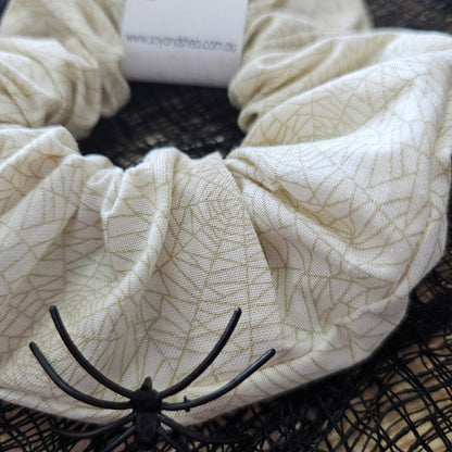 A detailed view of the Cobwebs Halloween  Scrunchie.  It shows a cream coloured scrunchie with a gold cobweb design all over