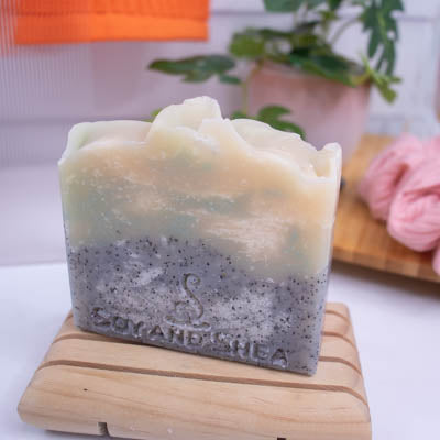 A bar of soap sits on a wooden soap dish on a bathroom counter top along with plant and scrunchie.  The Soap has two layers with the base being grey with black flecks and the top is cream with green swirls.