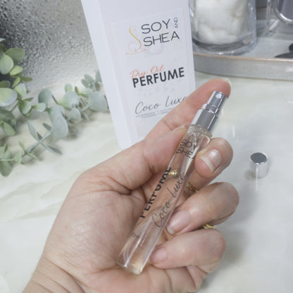 A hand holds a tall, narrow, clear perfume bottle that has a silver atomiser.  Behind is the packaging of a white box labelled as Dry Oil Perfume  on a bathroom countertop