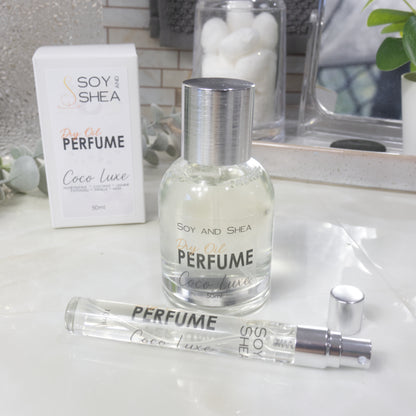 A selection of perfume bottles are presented on a bathroom countertop.  To the front is a clear, narrow, tall bottle labeled as 10ml with a silver cap and behind is a clear round, short bottle with silver lid labelled as 50ml.  Behind sits a white box with label identifying as Dry Oil Perfume Coco Luxe.