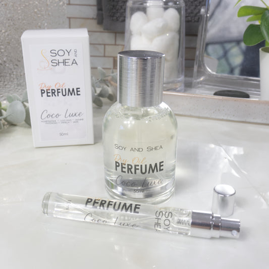 A selection of perfume bottles are presented on a bathroom countertop.  To the front is a clear, narrow, tall bottle labeled as 10ml with a silver cap and behind is a clear round, short bottle with silver lid labelled as 50ml.  Behind sits a white box with label identifying as Dry Oil Perfume Coco Luxe.