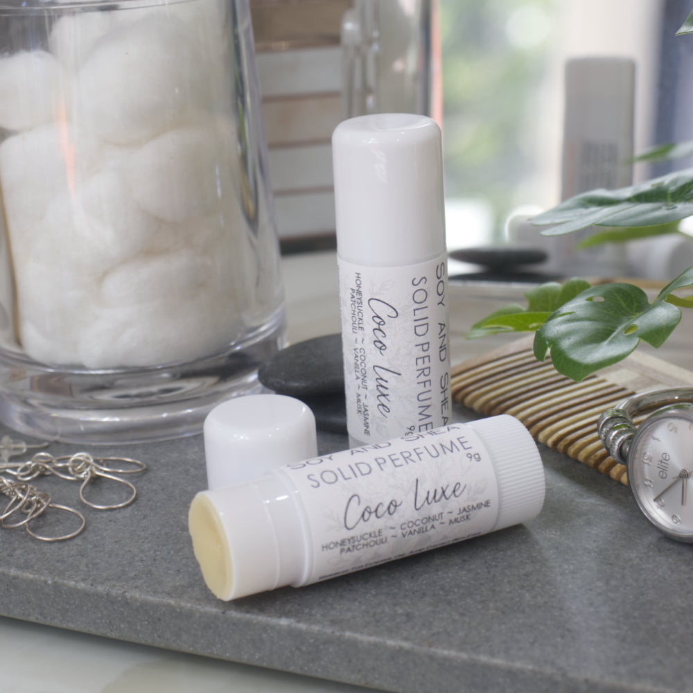 Two white tubes of Solid Perfume are displayed on a grey stone tray along with silver earrings, watch and greenery.   One tube lays flat displaying the fragrance notes while another tube stands upright behind it. The tube to the front has the lid removed revealing a beige balm.