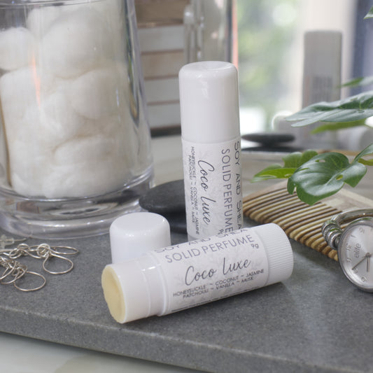Two white tubes of Solid Perfume are displayed on a grey stone tray along with silver earrings, watch and greenery.   One tube lays flat displaying the fragrance notes while another tube stands upright behind it. The tube to the front has the lid removed revealing a beige balm.