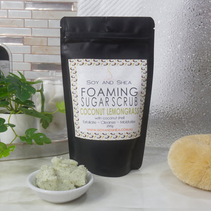 A black standup, ziplock pouch stands on a bathroom counter top by a shower screen with body brush and plant.  The bag features a product label identifying it as Foaming Sugar Scrub scented with Coconut Lemongrass.   To the front is a small white bowl with a sample of the pale green scrub which has small brown flecks of coconut shell