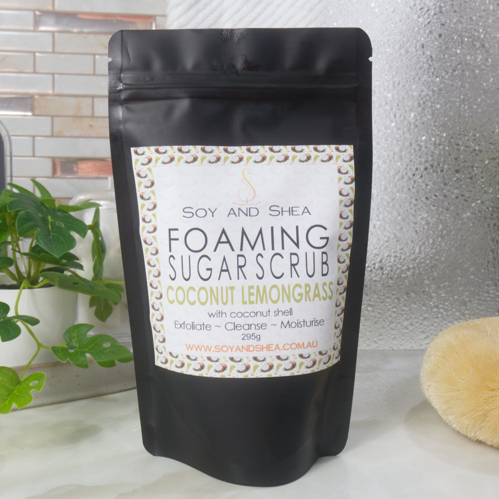 A black Pouch with Ziplock closure stands on a bathroom countertop surrounded by a plant, shower screen and body brush.  The pouch has a decorative label identifying it as Foaming Sugar Scrub Scented with Coconut Lemongrass