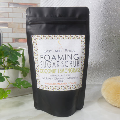 A black Pouch with Ziplock closure stands on a bathroom countertop surrounded by a plant, shower screen and body brush.  The pouch has a decorative label identifying it as Foaming Sugar Scrub Scented with Coconut Lemongrass