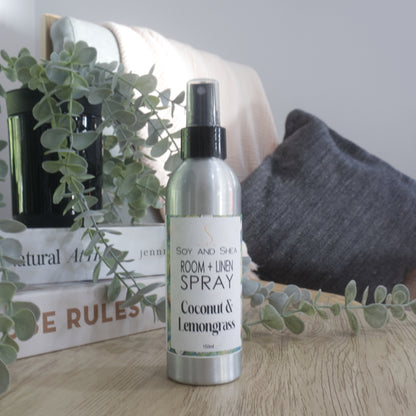 A silver room and linen spray bottle with a black spray top, labeled 'Soy and Shea Room + Linen Spray Coconut & Lemongrass', standing on a wooden surface with decorative books, a plant, and a cushion in the background.