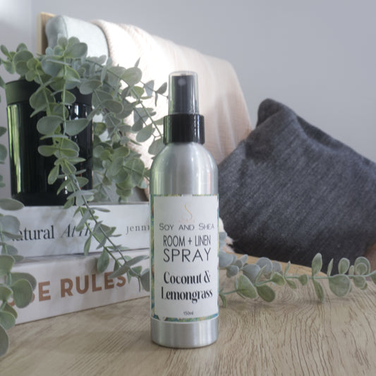A silver room and linen spray bottle with a black spray top, labeled 'Soy and Shea Room + Linen Spray Coconut & Lemongrass', standing on a wooden surface with decorative books, a plant, and a cushion in the background.