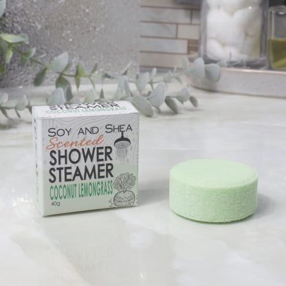 A boxed scented shower steamer and the steamer itself on a bathroom counter. The box reads 'Soy and Shea Scented Shower Steamer Coconut Lemongrass', and the round pale green steamer sits beside it.