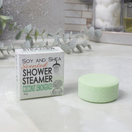 A boxed scented shower steamer and the steamer itself on a bathroom counter. The box reads 'Soy and Shea Scented Shower Steamer Coconut Lemongrass', and the round pale green steamer sits beside it.