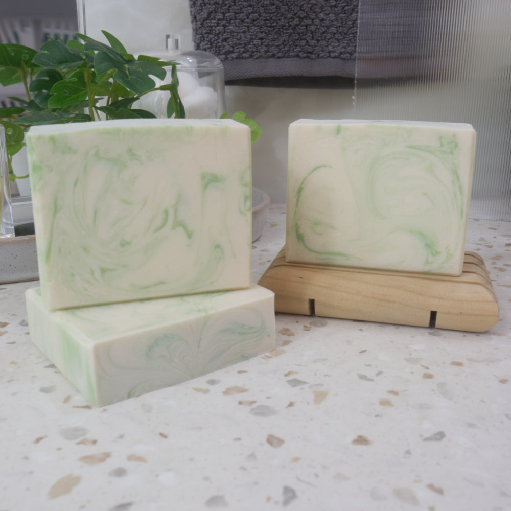 A selection of three soap bars are displayed on a bathroom counter top.  One soap lays flat on the left side showing a flat top soap.  Another stands upright on top of this and the right is a third soap presented on a wood soap dish.  The soaps are white with green swirls/