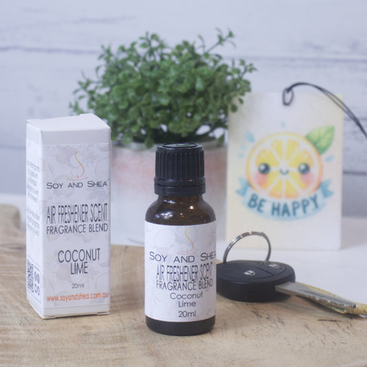 A small amber bottle with black lid labelled AIR FRESHENER SCENT stands on a wooden surface next to the box packaging.  In the background is a car key, air freshener and greenery.