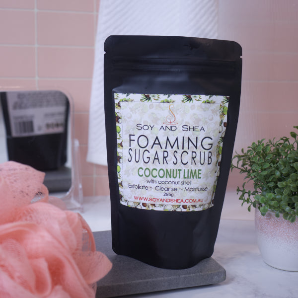 A black zip lock pouch stands upright on a stone tray in front of blurred bathroom background with peach tiles.   The pouch, which stands between a potted plant and a loofah, has a label that reads FOAMING SUGAR SCRUB COCONUT LIME and features images of coconut halfs against a white background