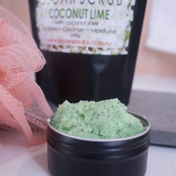 A zoomed in image of a black tin filled with green sugar scrub with brown flecks sitting on a white bench with the blurred image of the packaging in the background.