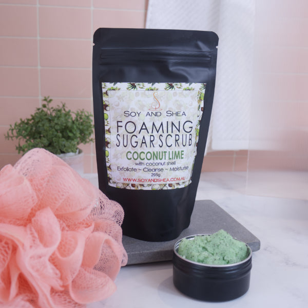 A black zip lock pouch stands up-right on a grey stone tray in front of pink tiled wall.  The Label reads  FOAMING SUGAR SCRUB COCONUT LIME against a white background covered with coconut halves.  In front is a small black tin overflowing with a green scrub with brown flecks.