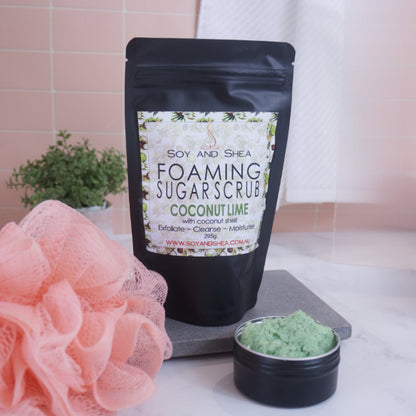 A black zip lock pouch stands up-right on a grey stone tray in front of pink tiled wall.  The Label reads  FOAMING SUGAR SCRUB COCONUT LIME against a white background covered with coconut halves.  In front is a small black tin overflowing with a green scrub with brown flecks.