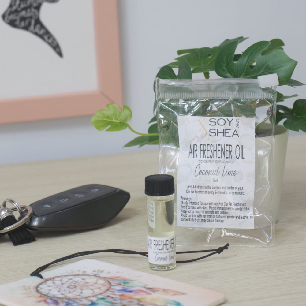A small clear bottle with black cap of Soy's Shea Air Freshener Oil next to a clear resealable pouch, displayed on a light wood tabletop with a car key, a felt air freshener, and a potted plant in the background.  