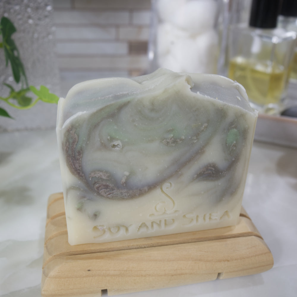 A close up image of a bar of soap standong on a wooden soap dish on a bathroom counter top along with blurred background.   The soap has a cream base colour with brown and light green swirls throughout.