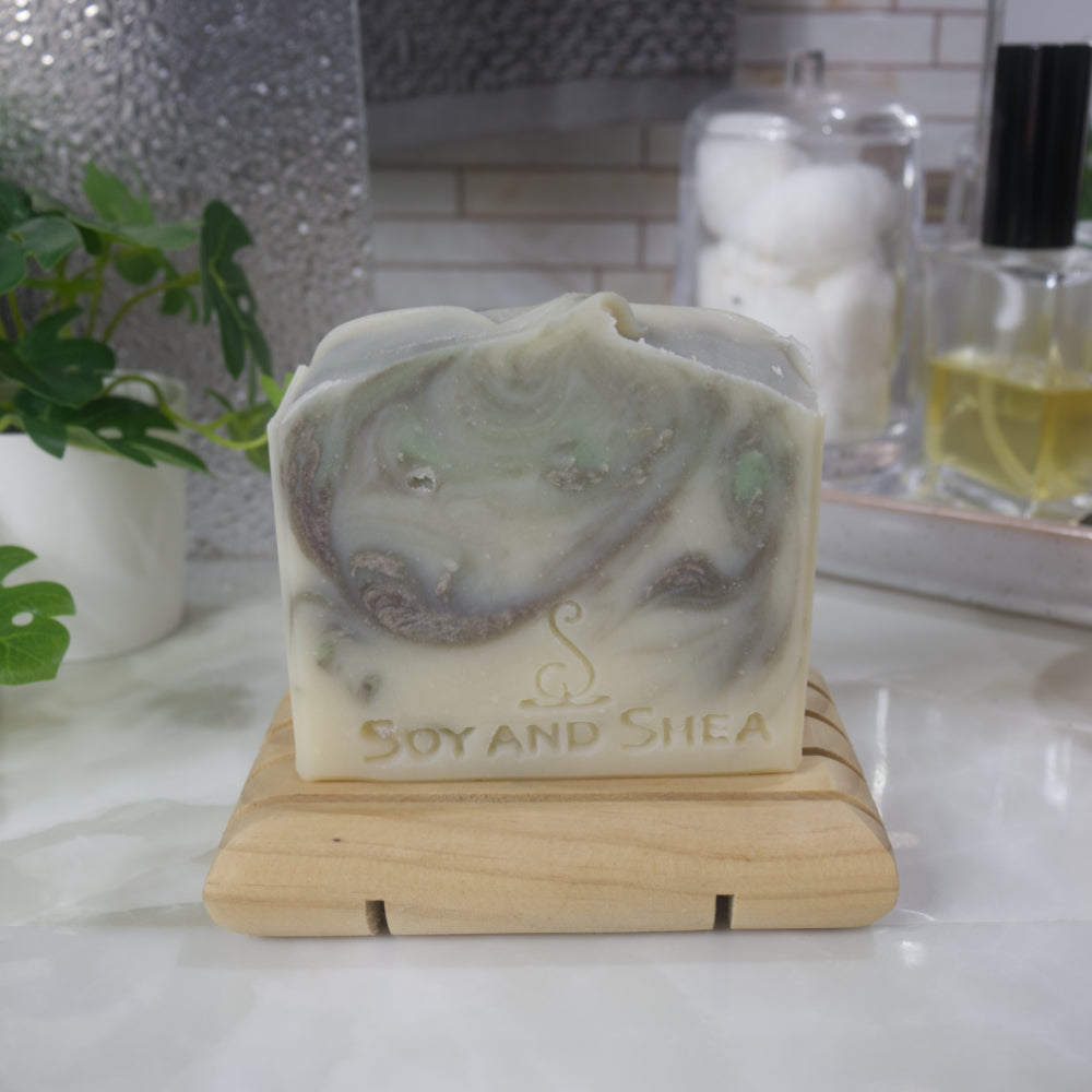 A bar of soap stands on a wooden soap dish on a bathroom counter top along with a plant, a jar of cotton balls and bottle of perfume.   The soap has a cream base colour with brown and light green swirls throughout.