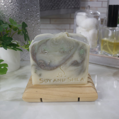 A bar of soap stands on a wooden soap dish on a bathroom counter top along with a plant, a jar of cotton balls and bottle of perfume.   The soap has a cream base colour with brown and light green swirls throughout.