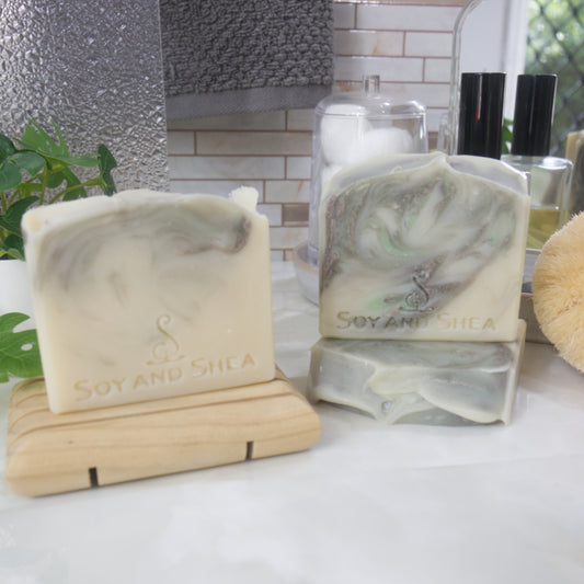 Three soap bars sit on a bathtoom countertop along with a perfume bottle, Jar of cotton balls and a plant.   One soap stands on a wooden soap rack to the left while another lies on its side to the right with a third standing on it.   The soaps have a cream base with brown and light green swirled through them.  Each soap has a slight variation in design.