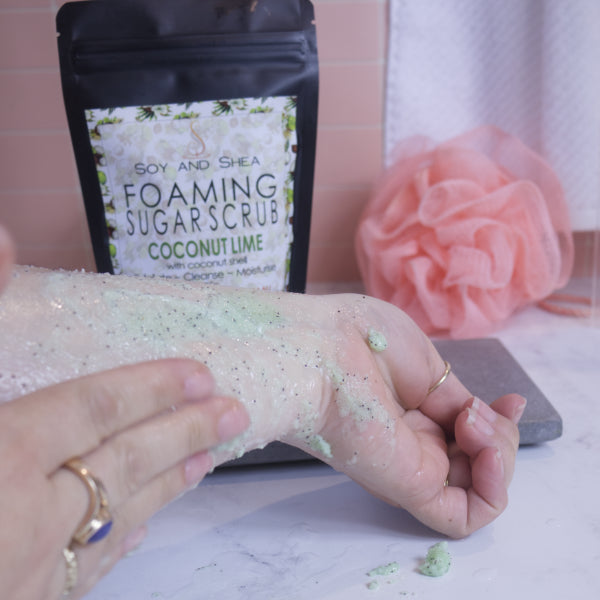 A hand applies a pale green scrub with brown flecks to the back of the other hand.  In the background sits a black pouch of the scrub with a label featuring coconuts, against a tile background with white towel and peach coloured loofah